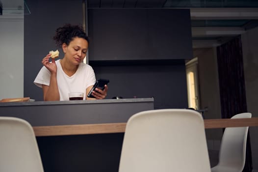 Confident multi ethnic woman 40s, using smartphone, checking social media content, browsing web, scrolling newsfeed, standing at kitchen counter with a sandwich in hand, enjoying her breakfast at home