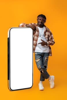 Smiling african man standing near huge smartphone with blank screen on yellow studio background, copy space