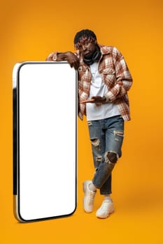 Smiling african man standing near huge smartphone with blank screen on yellow studio background, copy space