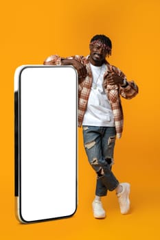 Smiling african man standing near huge smartphone with blank screen on yellow studio background, copy space