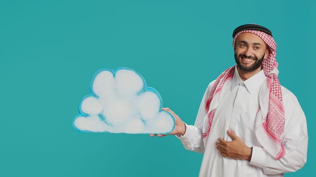 Middle eastern guy presents cloud board in studio, holding isolated billboard cutout in natural shape. Person wearing traditional arab islamic clothing and headscarf doing new web advertisement.