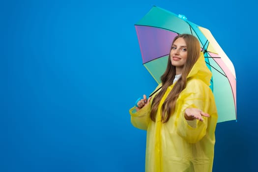 Young woman in yellow raincoat with rainbow umbrella against blue background in studio, close up