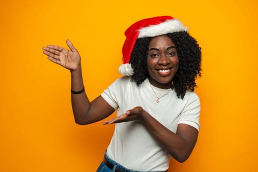 Happy excited african woman in santa claus hat pointing to copy space on yellow background in studio