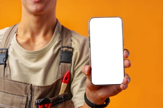 Portrait of handyman in uniform showing blank mobile phone on yellow background in studio