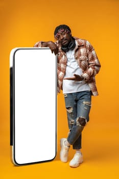 Smiling african man standing near huge smartphone with blank screen on yellow studio background, copy space