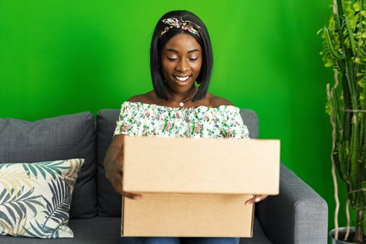 African young woman sit on couch at home unpack cardboard box with online purchase, close up