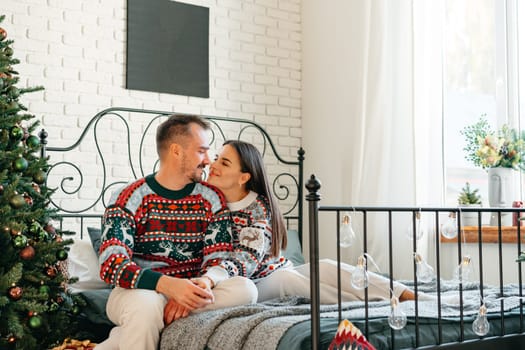 Young woman and man sitting on bed in sweaters and hugging near Christmas tree close up