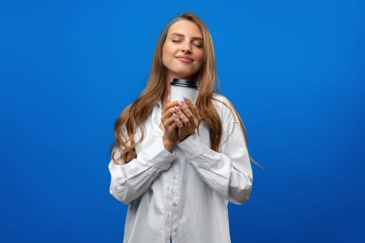 Beautiful attractive young lady holding takeaway paper coffee cup against blue background in studio, close up