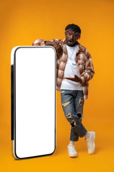 Smiling african man standing near huge smartphone with blank screen on yellow studio background, copy space