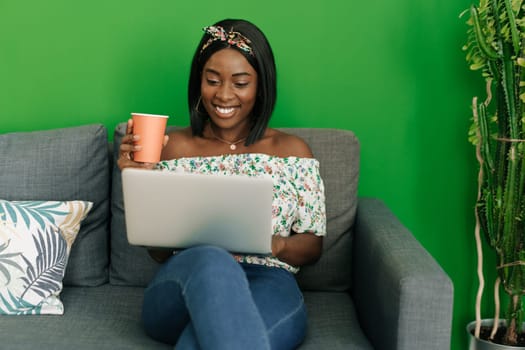 Young african woman working on laptop computer at home sitting on sofa close up