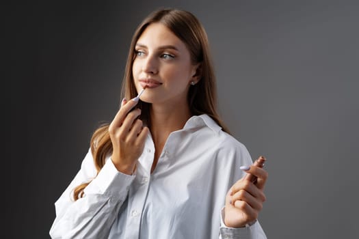 Cute young woman applying lip gloss against gray background in studio