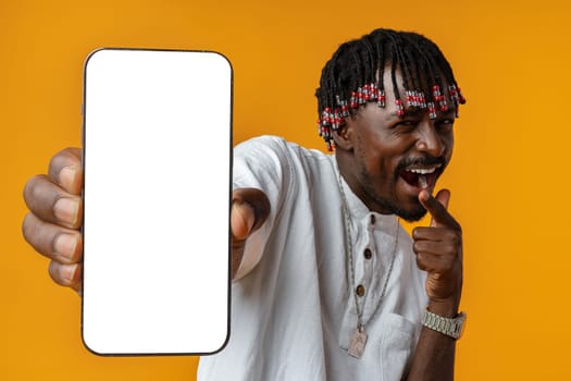 Excited young african man holding smartphone with white blank screen in hand on yellow studio background, copy space