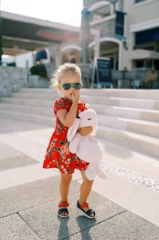 Little girl walks down the street past the steps of a building, picking her nose. High quality photo