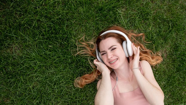 Top view of a young red-haired woman lying on the grass and listening to music on headphones