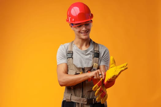 Young construction worker in helmet and uniform posing on yellow background in studio, close up