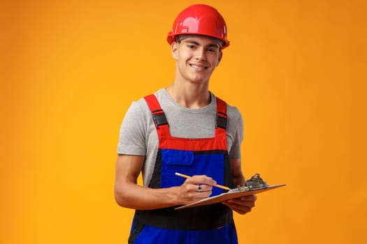 Young construction worker wearing hardhat and holding a clipboard on yellow background in studio, close up