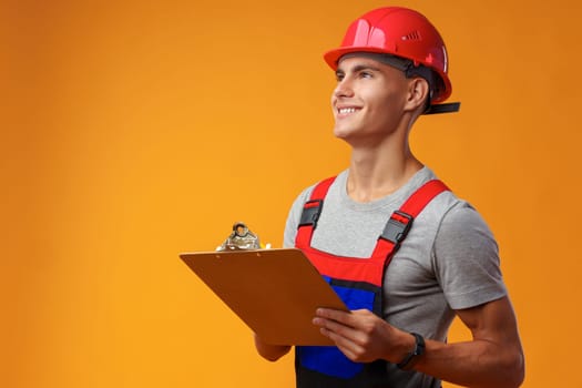 Young construction worker wearing hardhat and holding a clipboard on yellow background in studio, close up