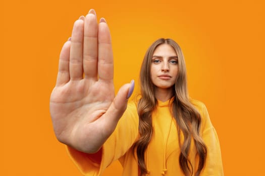 Young woman standing with outstretched arm showing stop sign against yellow background, close up