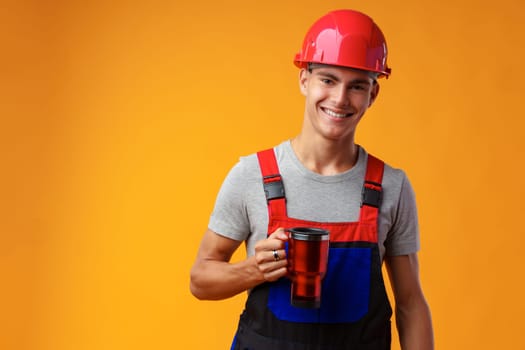 Young builder in uniform holding coffee cup on yellow background in studio