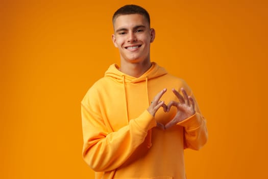 Portrait of young smiling man showing heart sign on yellow background in studio