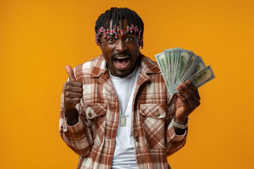Young happy rich african man in casual shirt holding dollar bills against yellow background close up