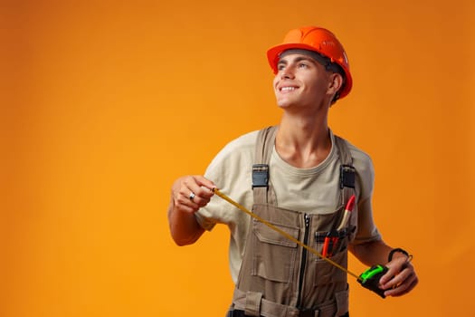 Young male builder holding measuring roulette against yellow background in studio