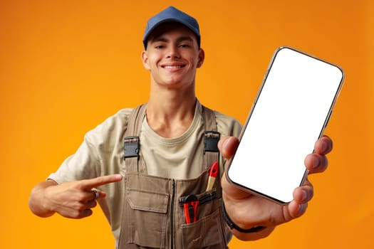 Portrait of handyman in uniform showing blank mobile phone on yellow background in studio