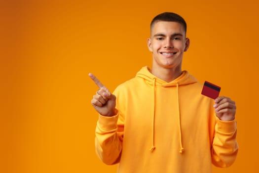 Handsome young man holding credit card on yellow color background in studio