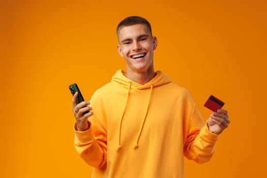 Young man holding credit card and smartphone against yellow background close up