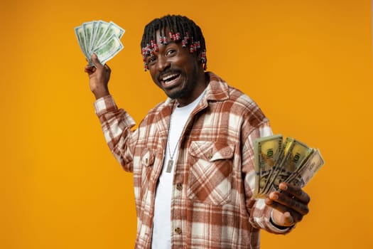 Young happy rich african man in casual shirt holding dollar bills against yellow background close up