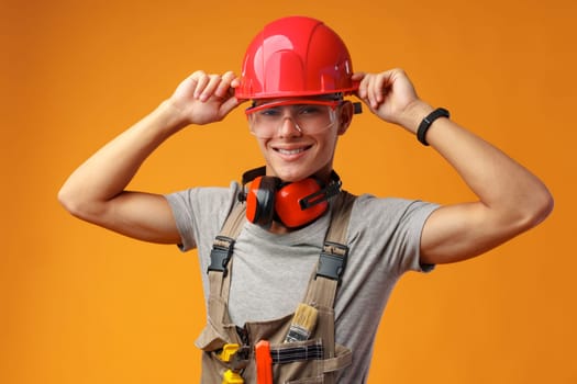 Young construction worker in helmet and uniform posing on yellow background in studio, close up