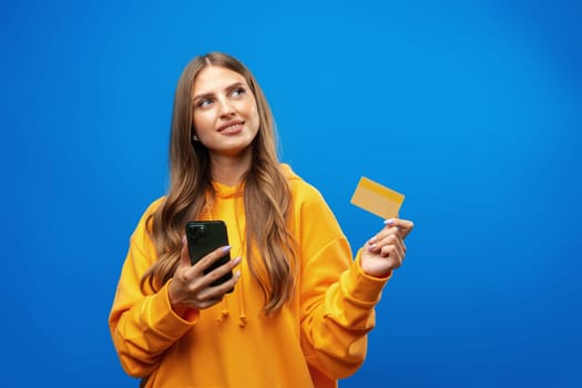 Portrait of a young blonde woman showing plastic credit card while holding mobile phone over blue background, close up