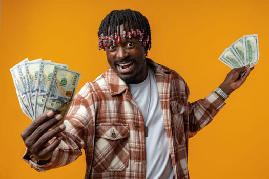 Young happy rich african man in casual shirt holding dollar bills against yellow background close up