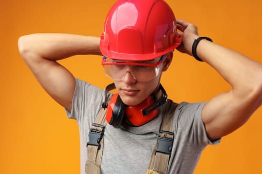 Young construction worker in helmet and uniform posing on yellow background in studio, close up