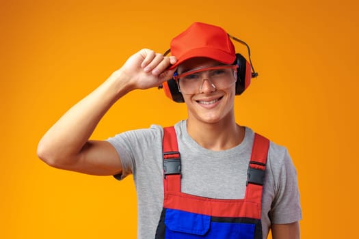 Young construction worker in helmet and uniform posing on yellow background in studio, close up