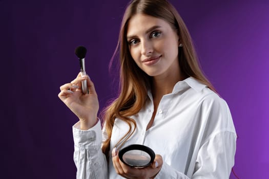 Young woman with makeup holding powder brush against purple background in studio