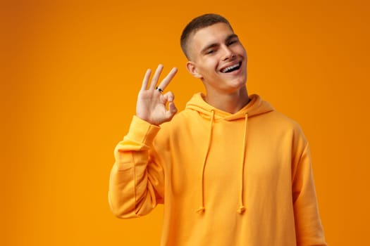 Happy young man gesturing OK sign against yellow studio background close up