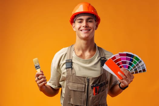 Handsome young worker in uniform holding paint samples palette on yellow background, close up