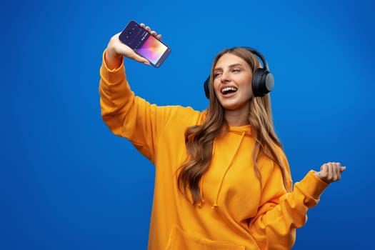 Smiling woman listening music in headphones and using smartphone over blue background in studio