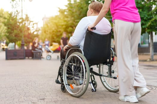 Back view of young woman helping mature woman in wheelchair strolling in the city