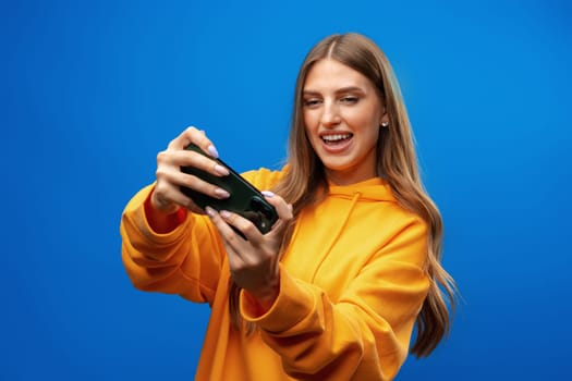 Excited young woman playing mobile video game on smartphone against blue background in studio