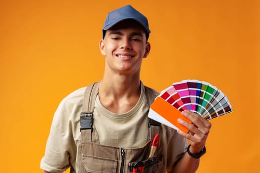 Handsome young worker in uniform holding paint samples palette on yellow background, close up