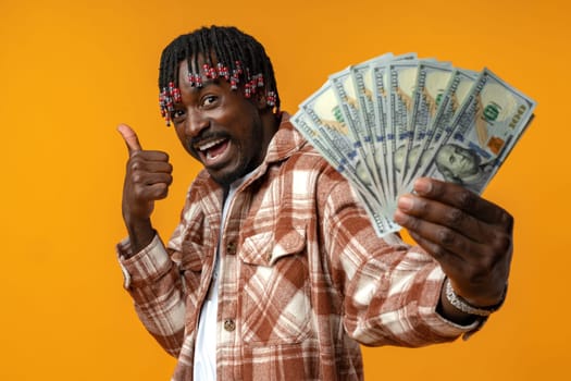 Young happy rich african man in casual shirt holding dollar bills against yellow background close up