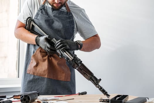 Close up of young man in apron disassembling a gun above the table in a workshop