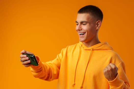 Portrait of excited young man playing online game on mobile phone on yellow background.in studio