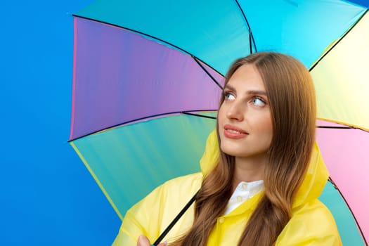 Young woman in yellow raincoat with rainbow umbrella against blue background in studio, close up