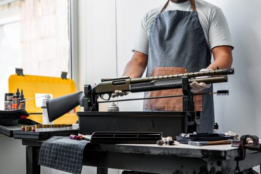 Close up of young man in apron disassembling a gun above the table in a workshop