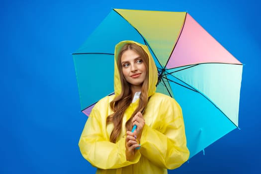 Young woman in yellow raincoat with rainbow umbrella against blue background in studio, close up