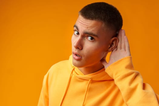 Interested young man hear something while keeping hand at his ear on yellow background in studio