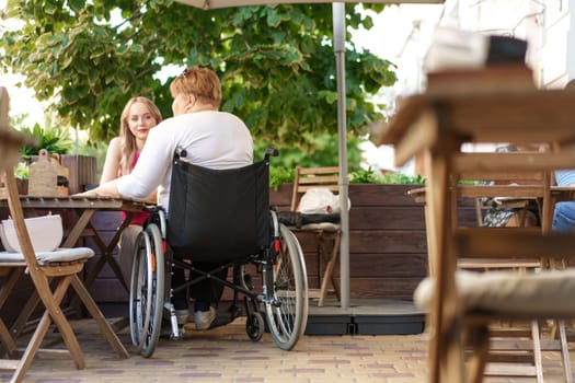 Mother with disability in wheelchair talking to her daughter while sitting at the table in cafe in the street